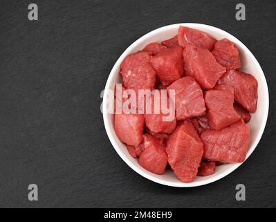 Rohes gewürfeltes Rindfleisch in weißer Schüssel auf schwarzem Schieferhintergrund, Draufsicht Stockfoto