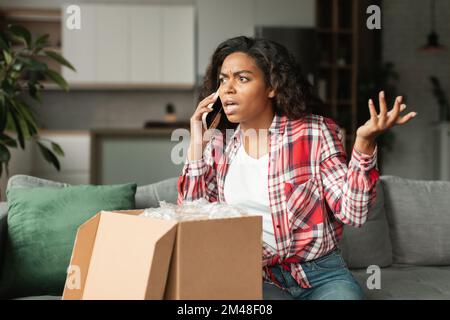 Schockierte, wütende Millenial Black Lady Anruf per Telefon, streitet mit dem Lieferservice im Wohnzimmer Stockfoto