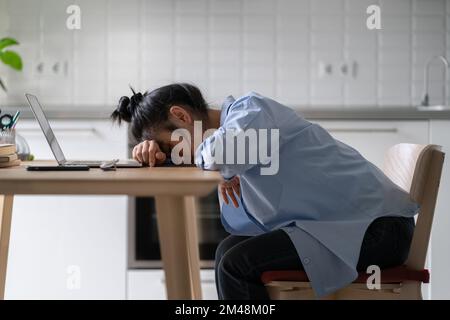 Müde, erschöpfte Freiberuflerin in Asien schläft, während sie von zu Hause aus arbeitet und auf dem Schreibtisch mit einem Laptop liegt Stockfoto
