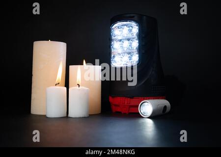 Elektrische Taschenlampen und brennende Kerzen auf schwarzem Hintergrund, alternative Licht- und Wärmequellen Stockfoto