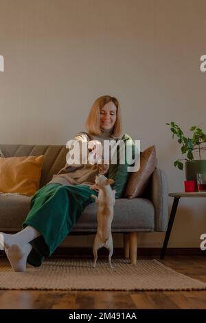 Eine Frau, die mit einem kleinen Hund spielt und auf dem Sofa sitzt. Das Privatleben. Stockfoto