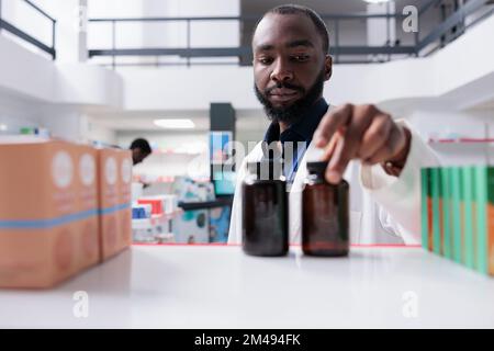 afroamerikanischer Apotheker, der Tablettenflaschen auf das Regal der Drogerie stellt, selektiver Fokus. Apotheke Verkäufer nimmt Medikamentenpakete aus der Nähe, verkauft Supplemente, Vorderansicht Stockfoto