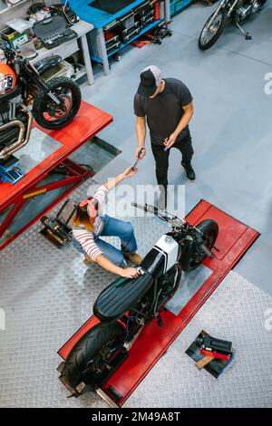Ein unerkannter Mechaniker, der einer Kollegin, die ein Motorrad auf Garaje repariert, Werkzeug gibt Stockfoto