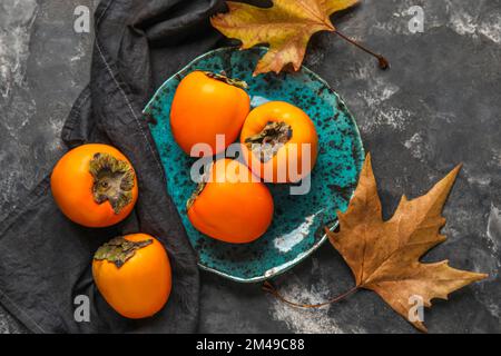 Teller mit reifen Persimonen und Herbstblättern auf dunklem Hintergrund Stockfoto