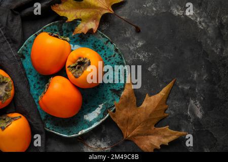 Teller mit reifen Persimonen und Herbstblättern auf dunklem Hintergrund Stockfoto