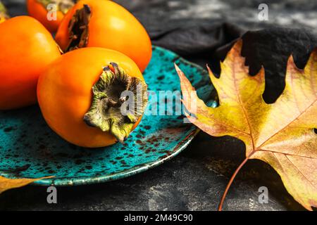 Teller mit reifen Persimonen und Herbstblättern auf dunklem Hintergrund, Nahaufnahme Stockfoto