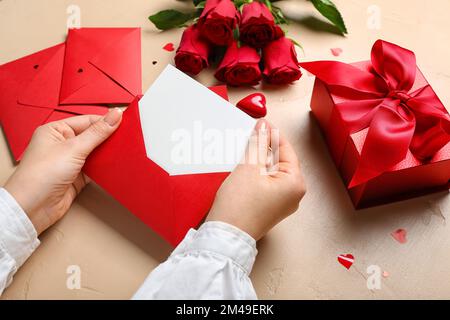 Frau mit leerem Brief, Rosen und Geschenk auf beigefarbenem Hintergrund. Valentinstag-Feier Stockfoto