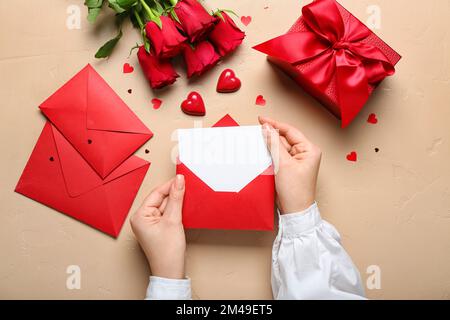 Frau mit leerem Brief, Rosen und Geschenk auf beigefarbenem Hintergrund. Valentinstag-Feier Stockfoto