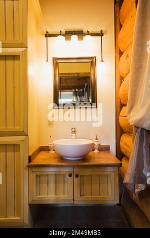 Weißes Waschbecken auf hölzernem Waschtisch im Hauptbadezimmer mit braunem Keramikfliesenboden im Blockhaus. Stockfoto