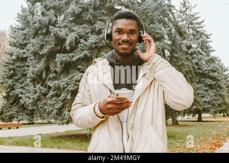 Ein hübscher Schwarzer mit kabellosen Kopfhörern, der Musik im Parkhintergrund hört. Lächelnder Afrikaner in lässiger Kleidung und weißen Kopfhörern Stockfoto