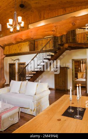 Langer hölzerner Esstisch mit beleuchteten Kerzen im Esszimmer und Sofa im Wohnzimmer im Blockhaus. Stockfoto