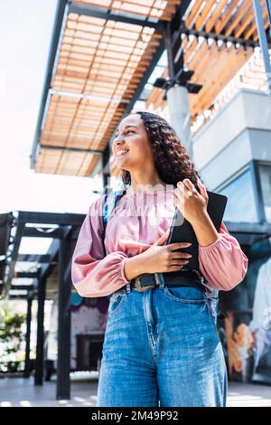 Schlichte, lächelnde hispanische Studentenfrau, die ihre Bücher hält und für den Unterricht bereit ist Stockfoto