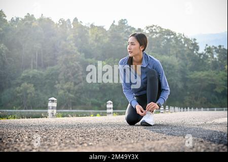 Entschlossene Asiatin in Sportbekleidung, die ihre Laufschuhe bindet und sich auf das Laufen auf der Straße vorbereitet. Konzept für einen aktiven gesunden Lebensstil Stockfoto