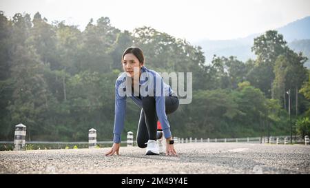 Entschlossene und fokussierte asiatische Läuferin in Sportbekleidung in der Startposition, die sich auf das Laufen auf der Straße vorbereitet. Stockfoto