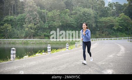 Eine glückliche und charmante Asiatin in aktiver Sportbekleidung, die beim Laufen auf der Straße Musik hört. Stockfoto