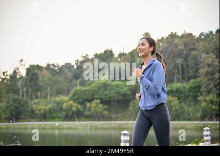 Charmante und glückliche Asiatin der Millennials in aktiver Sportbekleidung, die morgens gern im Park läuft und Musik hört. Stockfoto