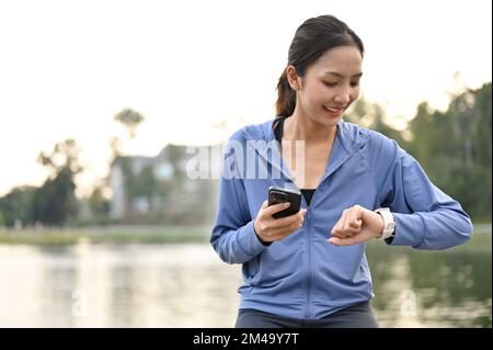 Fröhliche und sportlich fit sitzende Asiatin in Sportbekleidung hält ihr Handy und überprüft ihre Herzfrequenz und Laufmeilen auf ihrer Smartwatch. Stockfoto