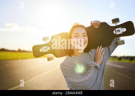 Fröhliches Skater-Mädchen, das auf die Kamera zugeht, Sonnenstrahlen scheinen auf die Linse. Junge Frau, die auf einem Kreuzer skatet, mit Longboard auf den Schultern Stockfoto