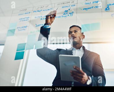 Es ist an der Zeit, diese Ideen aufzuwerten. Ein junger Geschäftsmann, der ein digitales Tablet benutzt, während er sich Notizen an eine Glaswand im Büro schreibt. Stockfoto