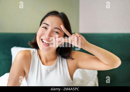 Positive asiatische Frau liegt im Bett, zeigt ein Friedenszeichen, genießt einen fröhlichen Morgen, wacht fröhlich auf, bleibt in ihrem Schlafzimmer Stockfoto