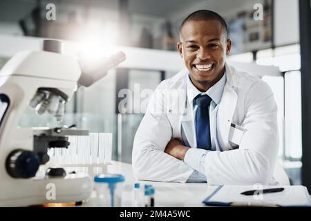 Alles im Namen der Wissenschaft. Abgeschnittenes Porträt eines gutaussehenden jungen Wissenschaftlers, der mit gefalteten Armen in seinem Forschungslabor sitzt. Stockfoto