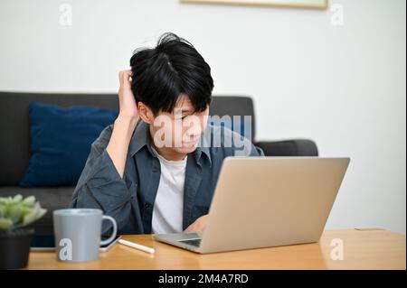 Verwirrter junger asiatischer Mann, der Probleme mit der Internetverbindung bei der Arbeit hat, Probleme mit seinem Laptop bekommt, und sich voreingenommen über sein Projekt Gedanken macht. Stockfoto