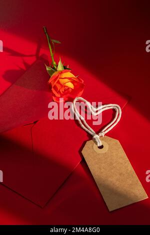 Schöne rote Rosen Blumen in postalischem roten Umschlag auf rotem Hintergrund, beige Papiernote und Seil in Herzform Kopieplatz für Text, Frühling, Grußkarte für Urlaub. Blumenlieferung. Zarte rote gelbe Rosen. Monochrome Minimal trendige Komposition. Romantische Blumen Stockfoto