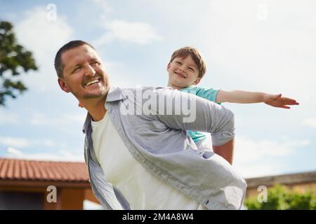 Wenn man daran glaubt, kann man es erreichen. Ein junger, gutaussehender Vater, der seinem süßen kleinen Sohn eine Huckepackfahrt im Hinterhof zuhause gibt. Stockfoto