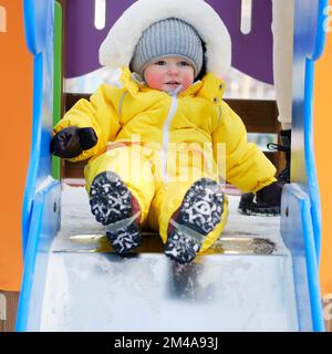 Ein kleiner Junge in einem gelben Schneeanzug fährt auf einem verschneiten Spielplatz eine Kinderrutsche hinunter. Ein Kind im Alter von einem Jahr, fünf Monate beim Spielen auf einem Winterspaziergang Stockfoto