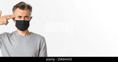 Konzept von covid-19, sozialer Distanzierung und Quarantäne. Nahaufnahme eines traurigen Mannes mittleren Alters in schwarzer medizinischer Maske, mit der Fingerpistole auf den Kopf zeigend und schauend Stockfoto