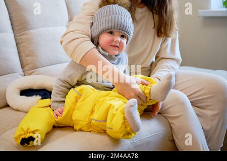 Das Kind trägt warme Winterkleidung für einen Spaziergang nach draußen. Weibliche Mutter zieht bei kaltem Wetter Klamotten an. Ein Kind im Alter von einem Jahr, fünf monate Stockfoto