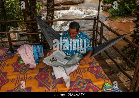 Kambodschanischer Mann liest Zeitung in der Hängematte, Kbal Chhay Wasserfall am Prek Tuk SAP River, Sihanoukville Provinz, Kambodscha. © Kraig Stockfoto