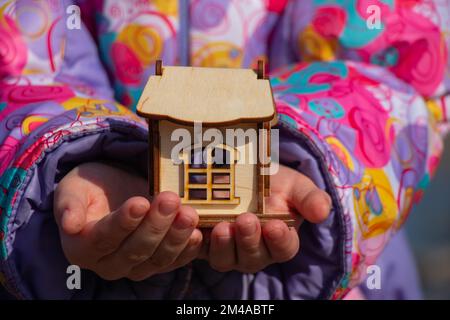 Die Kinderhand hält ein kleines Holzhaus auf einem verschwommenen Hintergrund Stockfoto