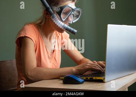 Ein ukrainisches Mädchen sitzt in einer Taucherbrille und Handsets an einem Laptop auf einem Tisch zu Hause und träumt vom Urlaub Stockfoto