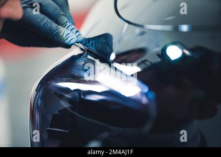 Nahaufnahme einer Person mit blauen Handschuhen, die sich hineinlehnt, um am Rücklicht eines schwarzen Autos in einer Garage zu arbeiten. Autoteile. Horizontale Indoor-Aufnahme. Hochwertiges Foto Stockfoto