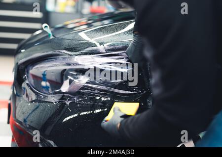 Nahaufnahme eines Mannes mit Handschuhen, der PPF-Lackschutzfolie auf dem Heck eines schwarzen Autos in einer Garage aufbringt. Autoteile. Horizontale Indoor-Aufnahme. Hochwertiges Foto Stockfoto