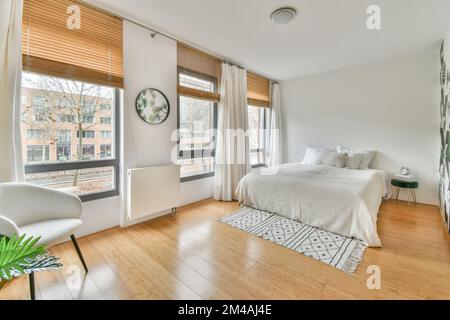 Ein geräumiges Schlafzimmer mit einem großen weißen Bett und einem großen Fenster daneben in einem gemütlichen Apartment Stockfoto