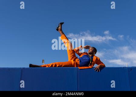 Tiefer Seitenblick des ganzen Körpers einer afroamerikanischen Frau mit kurzen Haaren im orangefarbenen Anzug und hohen Absätzen mit erhobenen Beinen auf blauer Wand und Spaß Stockfoto