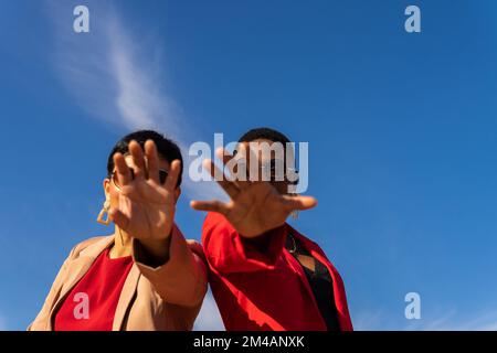 Ein kleiner Winkel von erwachsenen, multiethnischen Freundinnen mit kurzen dunklen Haaren in stilvollen Anzügen und einer Sonnenbrille, die beim Stehen die Hände in Richtung Kamera strecken Stockfoto