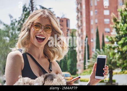 Fröhliche junge, stilvolle Millennials mit welligen blonden Haaren in trendigen Outfits und einer Sonnenbrille, die fröhlich schreit und in die Kamera schaut, während sie auf SM zeigt Stockfoto