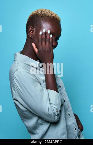 Schäbiges afroamerikanisches Weibchen in einem stilvollen Hemd mit kurzen blonden Haaren, die das Gesicht mit der Hand vor blauem Hintergrund bedecken Stockfoto
