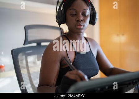 Junge afroamerikanische Frau, die Musik hört und mit dem Eingabestift auf dem Grafiktisch malt, während sie am Schreibtisch sitzt und an einem freiberuflichen Projekt arbeitet Stockfoto