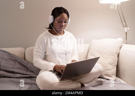 Junge afroamerikanische Frau in weißem Pyjama und Socken mit Kopfhörern, die auf einem Laptop Filme durchsucht, während sie im Wohnzimmer mit einem Kreuzbein auf dem Sofa sitzt Stockfoto