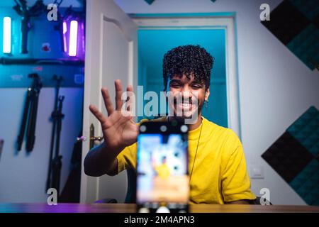 Fröhlicher junger ispanischer Mann mit lockigem Haar, das lächelt und der Hand winkt, während er am Schreibtisch hinter dem Smartphone sitzt und zu Hause Videos für einen Blog aufnimmt Stockfoto
