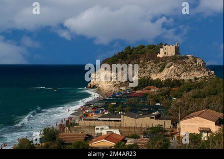 Urlauber paddeln im Tyrrhenischen Meer am Sandstrand unter dem Heiligtum Santa Maria dell’Isola, das nach Erdbeben wieder aufgebaut wurde und sich auf einem felsigen Vorsprung unterhalb der Klippenstadt Tropea auf den westlichen Kosten von Kalabrien in Süditalien befindet. Der Felsen, der einst eine Insel war, ist seit vielen Jahrhunderten eine Klosterstätte. Stockfoto