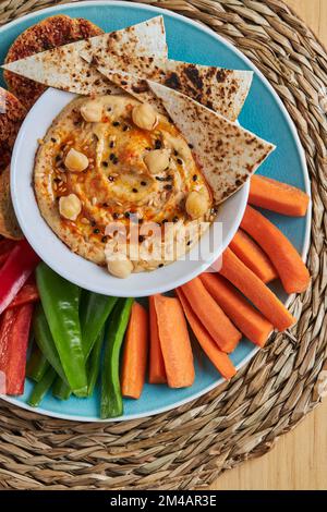 Blick von oben auf die Schüssel mit Hummus und Tortilla-Chips, umgeben von verschiedenen Gemüse und Croutons, serviert auf Korbmatte auf Holztisch Stockfoto