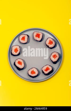Blick von oben auf eine Portion köstlicher Sushi-Brötchen mit Reis und Lachs in Nori verpackt, serviert auf einer grauen Keramikplatte auf gelber Oberfläche Stockfoto