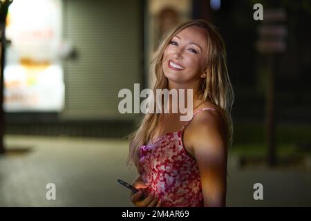 Glückliche Frau mittleren Alters in stilvollen Kleidern mit lächelndem Handy und Blick über die Schulter auf die Kamera auf verschwommenem Hintergrund einer dunklen Stadtstraße bei Nacht Stockfoto