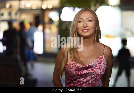 Glückliche junge Blondine, die nachts mit einem Lächeln auf einem unscharfen Straßenhintergrund in die Kamera schaut Stockfoto