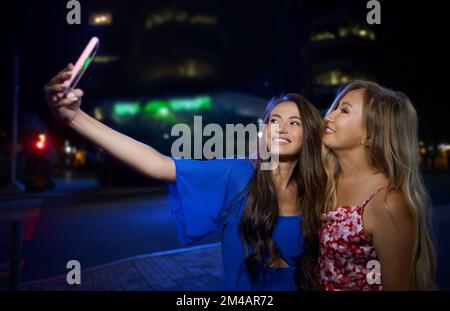 Fröhliche junge Brünette in stilvollem blauem Kleid, lächelnd und Selfie mit reifer mutter, während sie nachts auf der Straße der Stadt steht Stockfoto